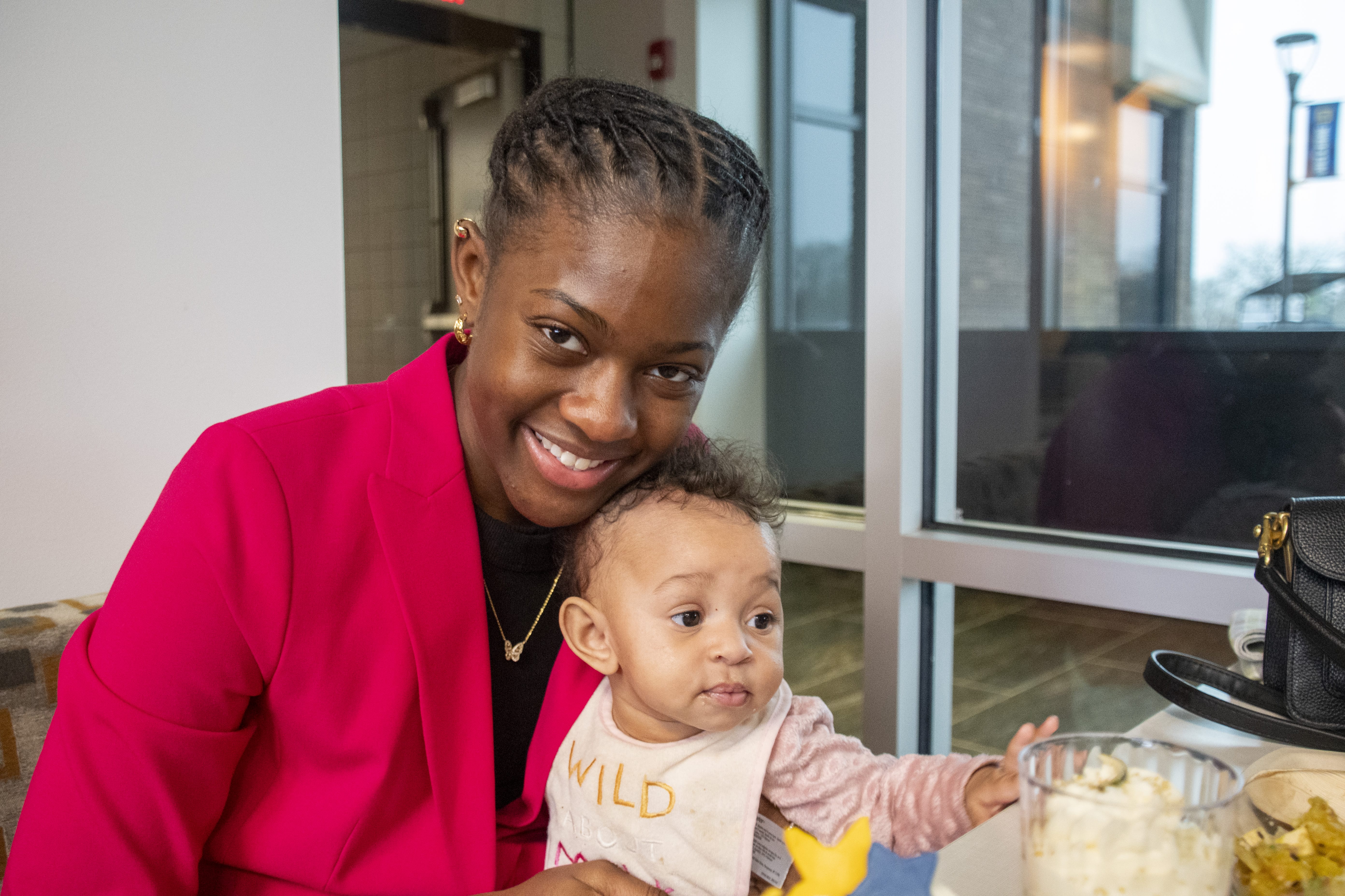 A smiling woman in a bright pink blazer sits next to a baby wearing a bib that says “WILD.” They are indoors by a large window, and a glass bowl of food is on the table in front of them.