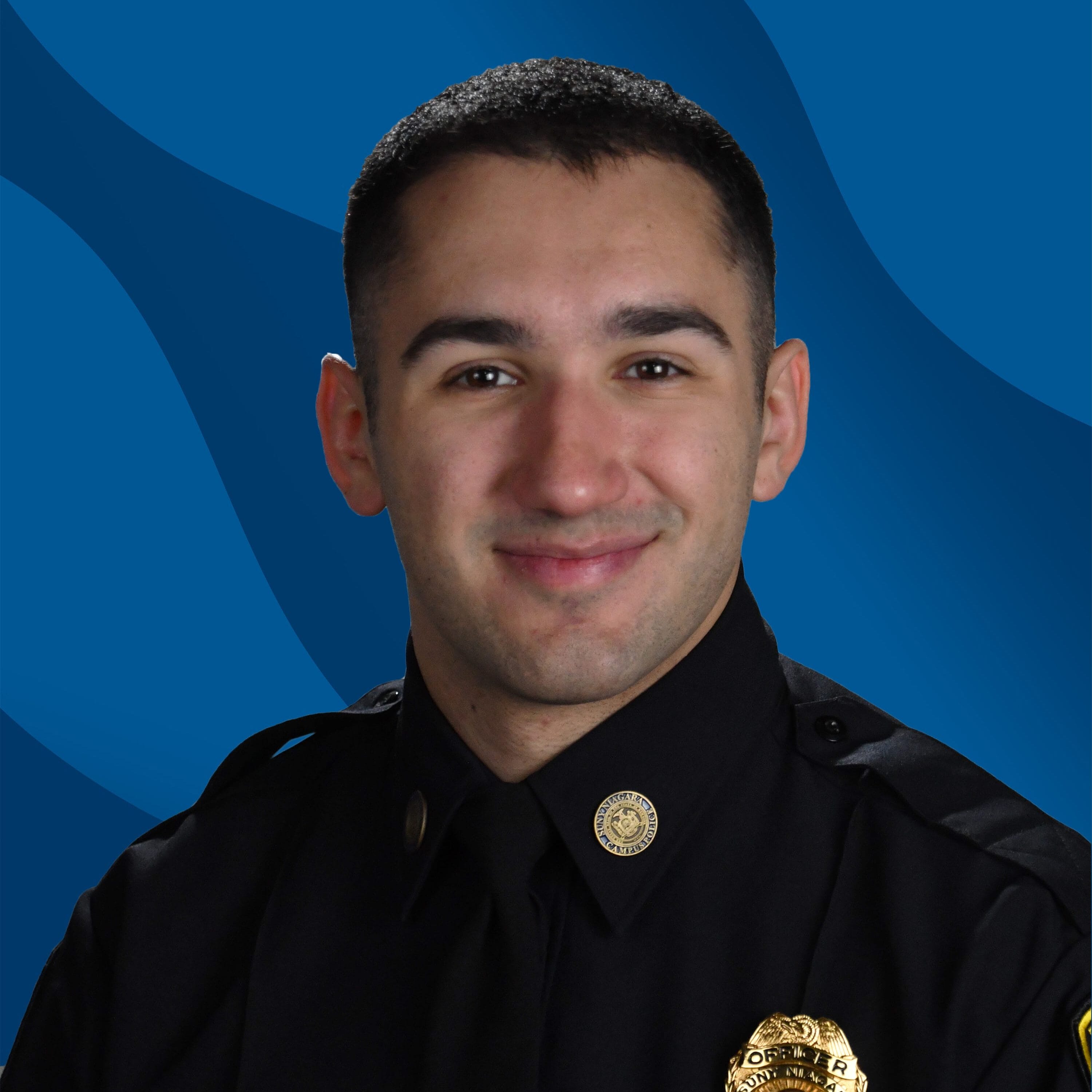 A young man in a black police uniform with badges and a tie smiles in front of a blue background with wavy patterns.