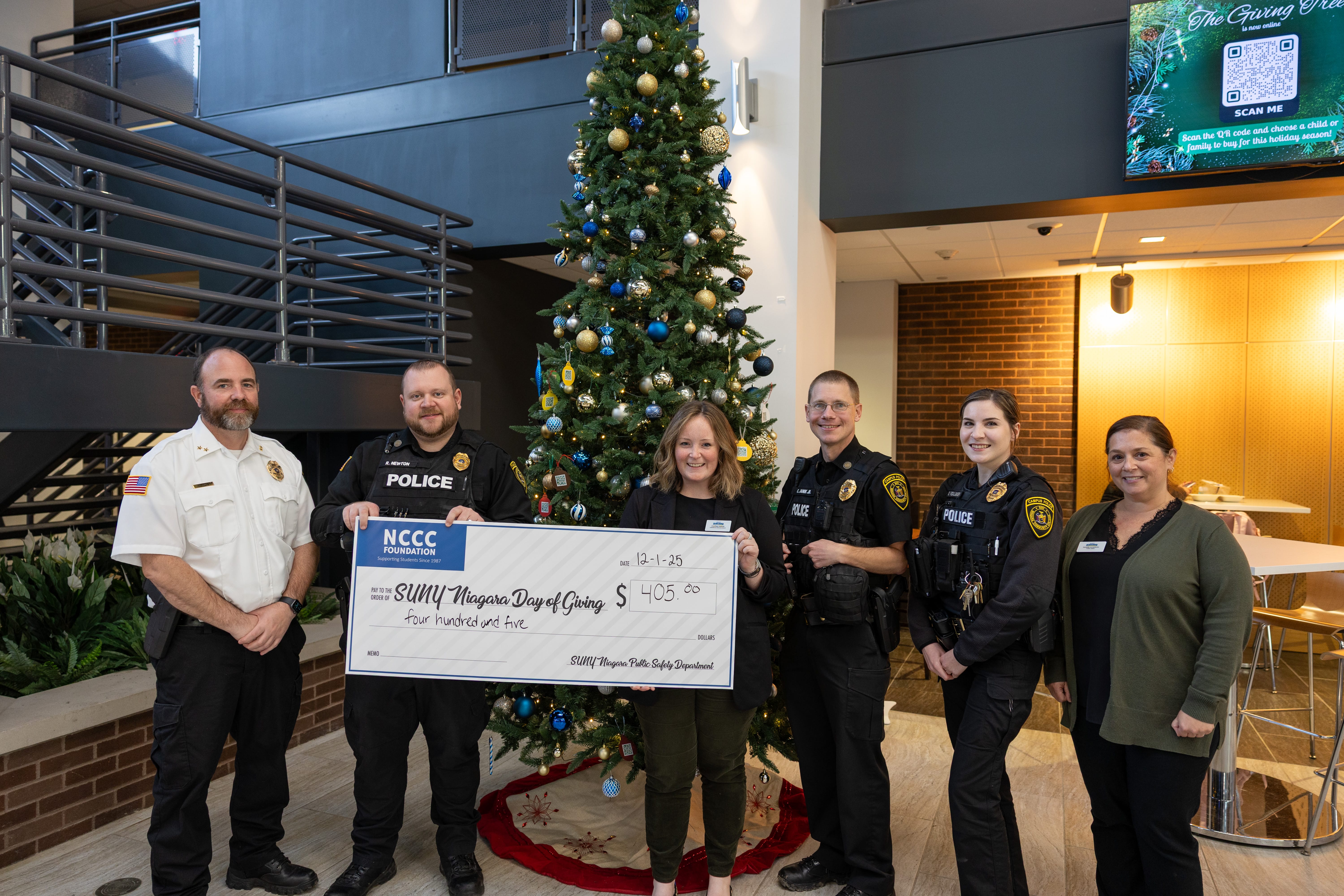 Six people, including police officers and staff, stand indoors by a decorated Christmas tree, smiling and holding a large ceremonial check for $405 made out to SUNY Niagara Daryl’s Closet.