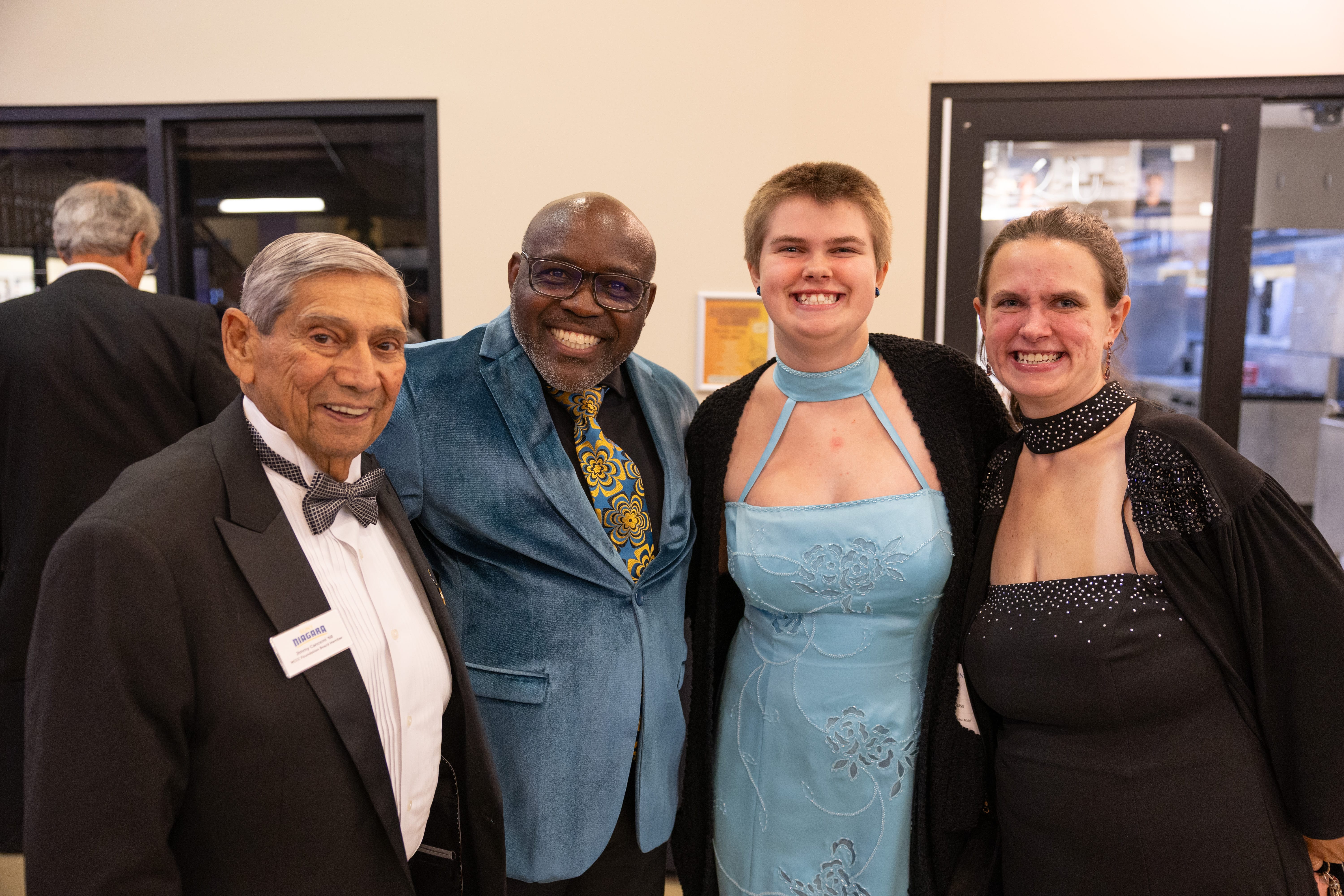 Four people dressed in formal attire smile for a group photo at an indoor event. Two men wear suits and bow ties, while two women wear elegant blue and black dresses. They appear happy and are standing close together.
