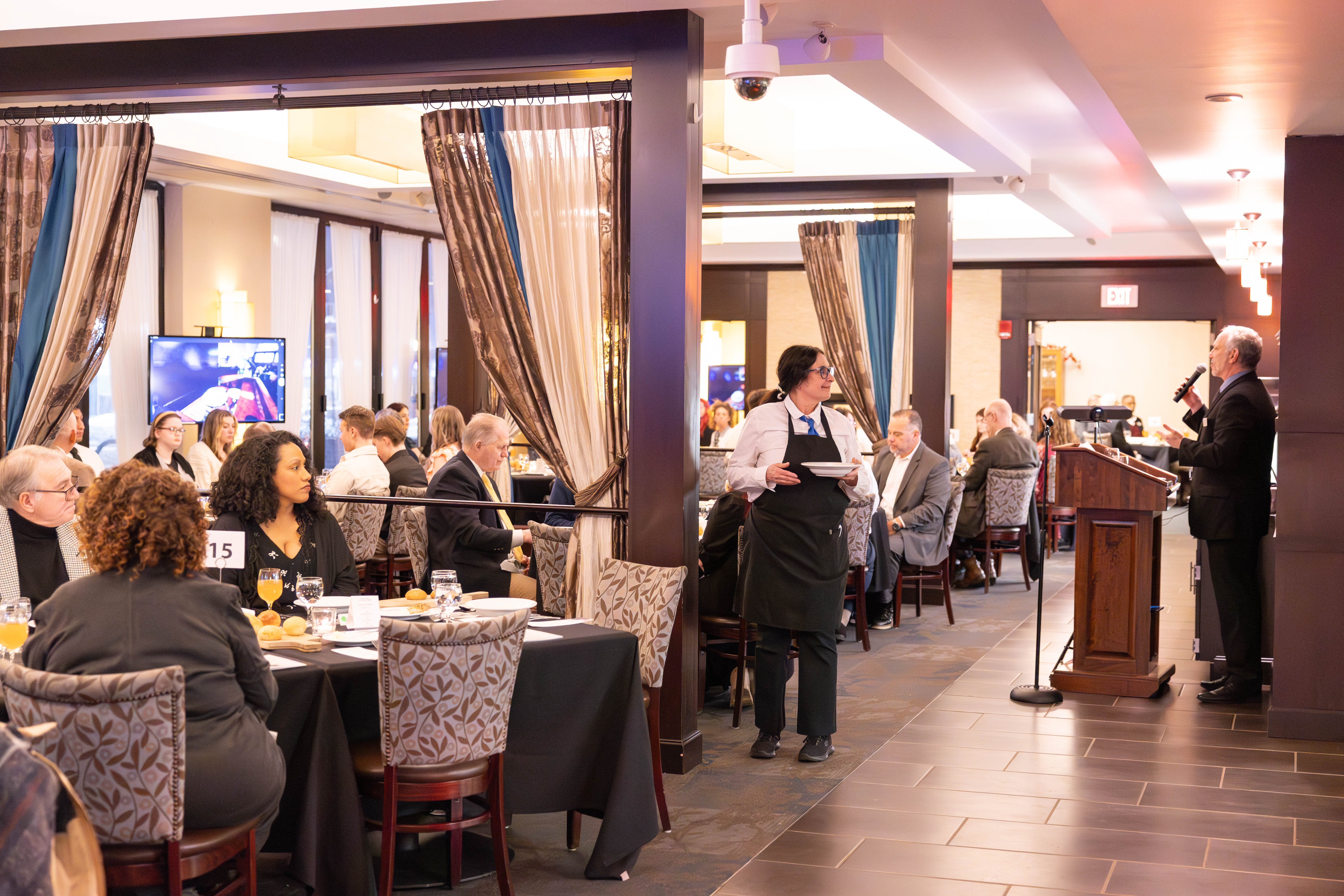A formal dining event with people seated at round tables, a waiter serving guests, and a speaker at a podium addressing the audience in a well-lit, elegant restaurant.