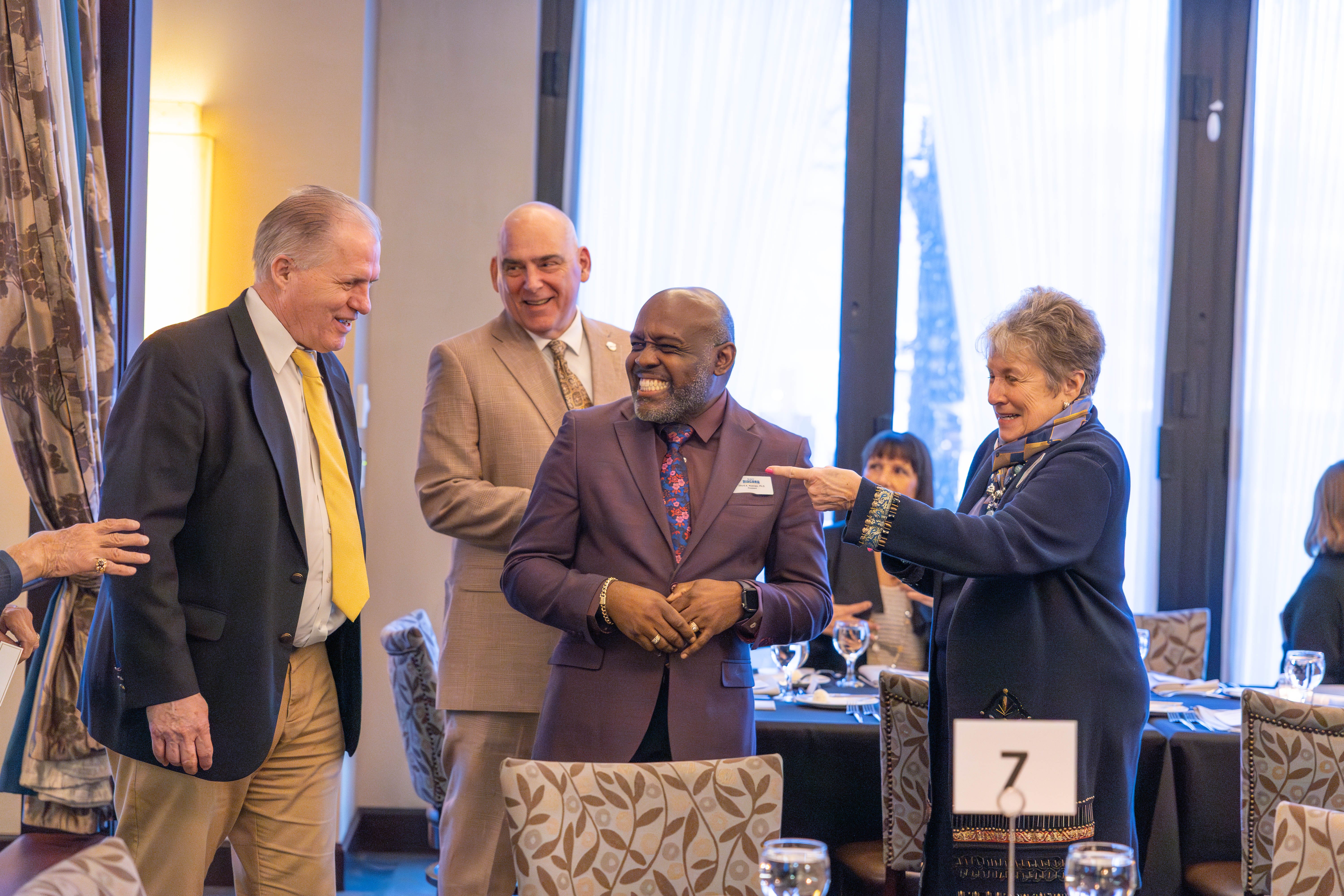 Four people stand and smile in a bright room set for a formal event; a woman gestures and laughs while three men, dressed in suits, smile. Other attendees sit at tables in the background. Table number 7 is visible.