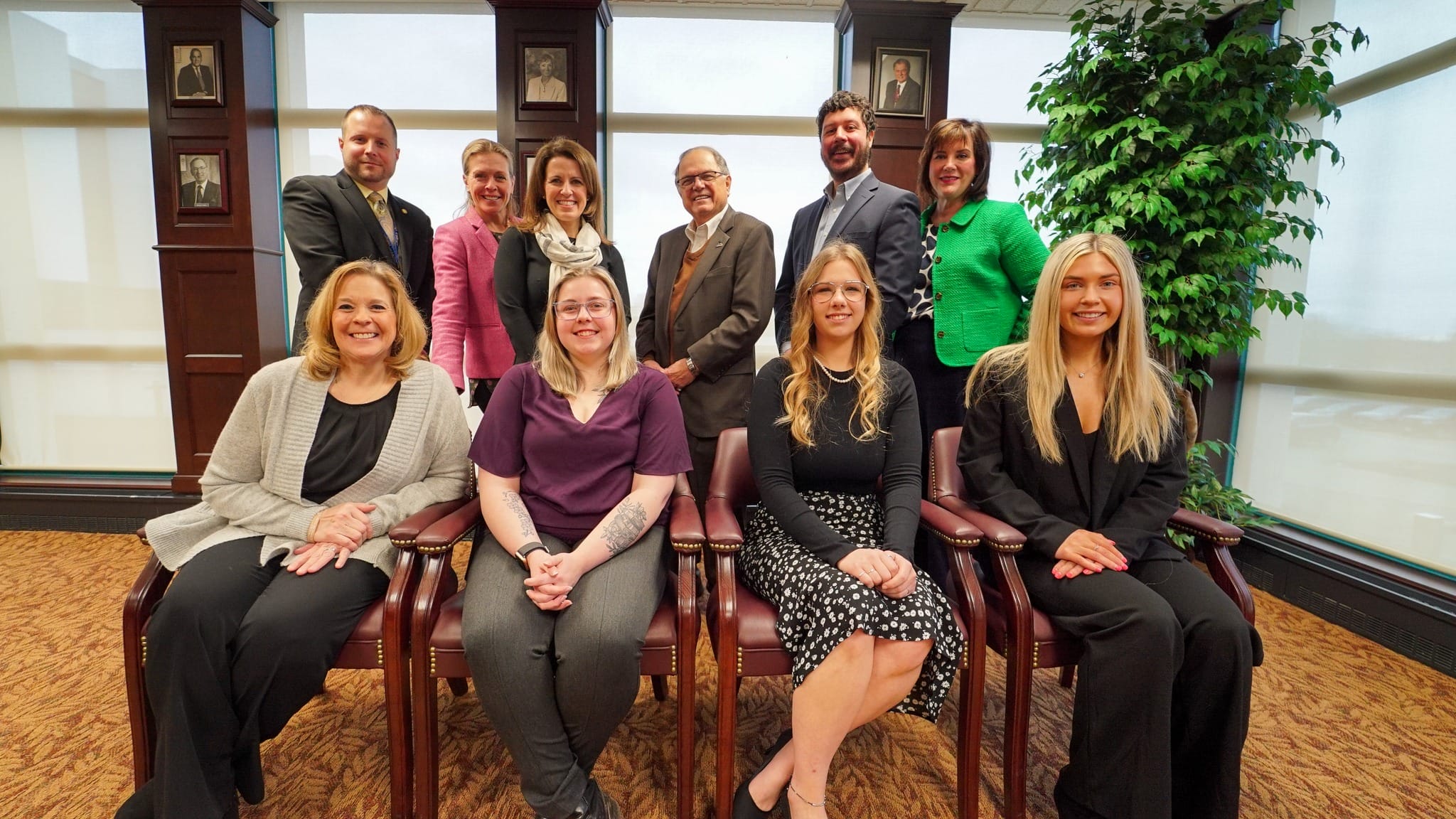 Ten adults, six women and four men, pose together indoors. Four people are seated in front, while six stand behind them. They are dressed in business or business casual attire, smiling for the photo.