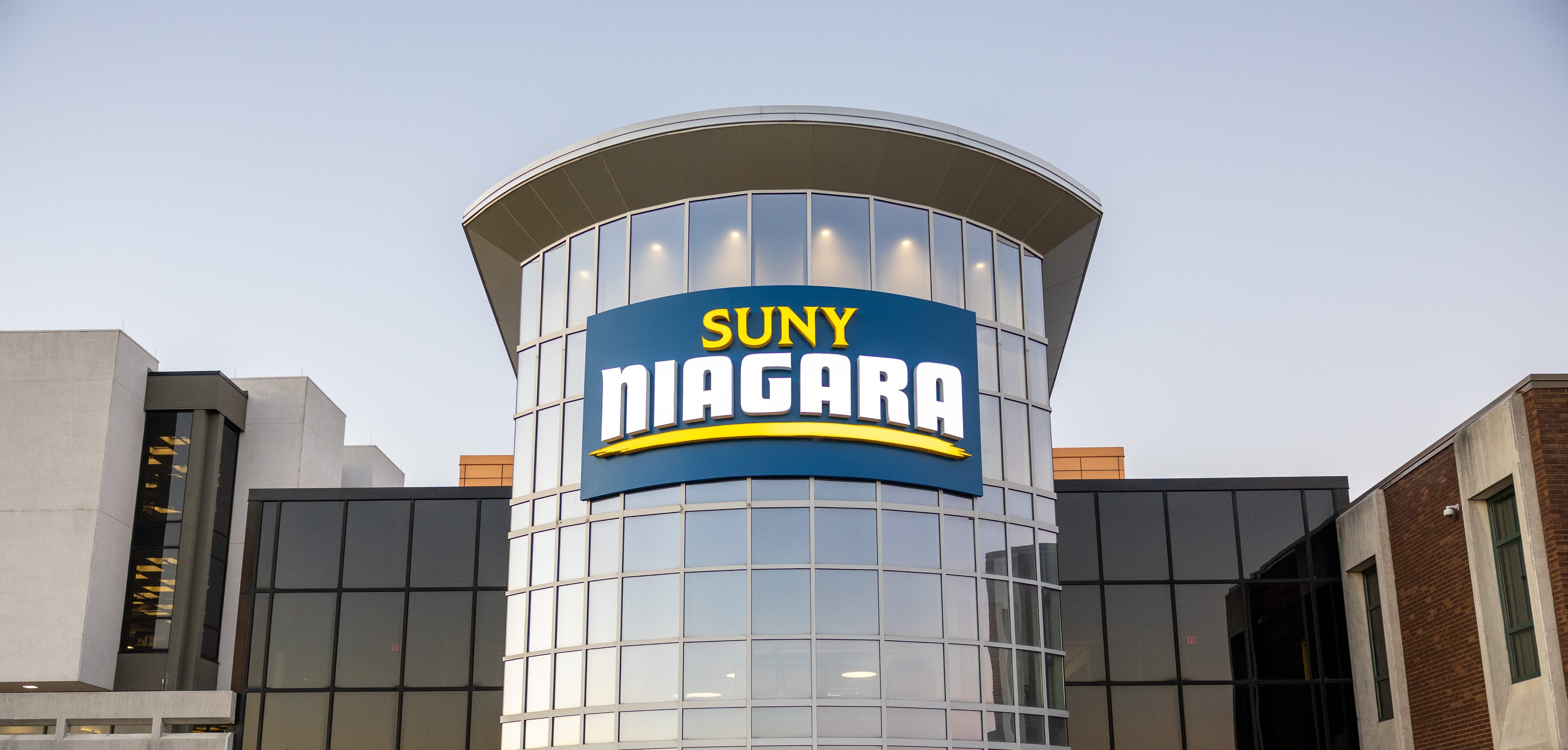 A modern glass building with a large sign reading SUNY NIAGARA above the entrance, flanked by brick and concrete sections, photographed at dusk.