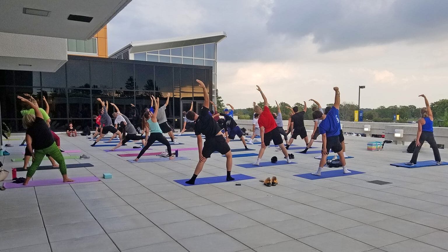 A group of people participate in an outdoor yoga class on a large patio, each on a yoga mat, stretching with one arm raised under a cloudy sky near a modern building.