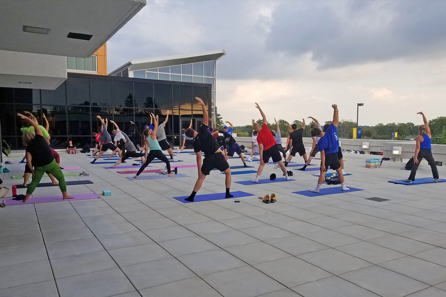 A group of people participate in an outdoor yoga on a large patio, each on a yoga mat, stretching with one arm raised under a cloudy sky near a modern building.