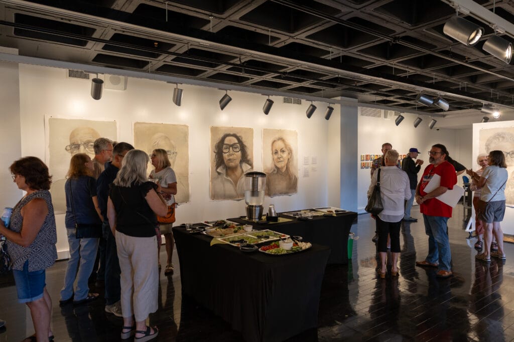 People gather in an art gallery, viewing large portrait drawings hung on white walls. Two tables with snacks and drinks are set up in the center. The atmosphere is lively and social.