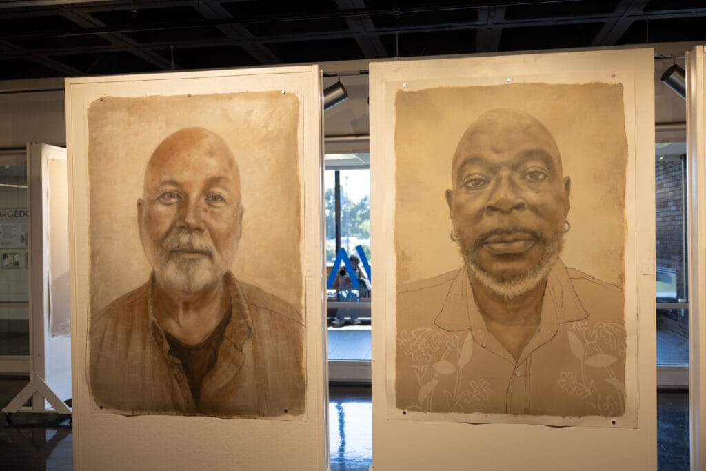Two portrait paintings are displayed side by side on white panels in a gallery. Each portrait depicts a man facing forward, with detailed, realistic features, set against a neutral background. Natural light enters from windows behind.