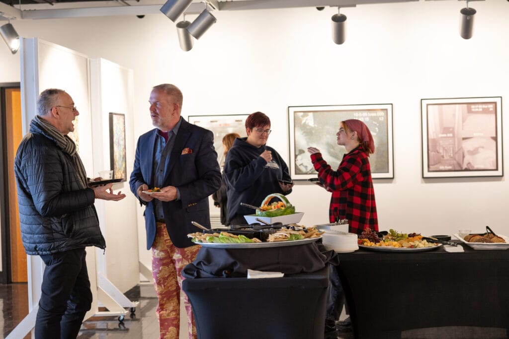 Five people stand and converse near buffet tables with assorted food platters at an indoor event. Artworks are displayed on the wall behind them. Two people are eating while talking, and one person wears a red checkered shirt.