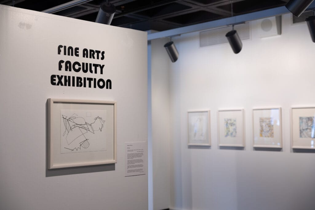 A gallery wall displays a sign reading Fine Arts Faculty Exhibition beside a framed abstract drawing; more framed artworks hang on the adjacent wall in a well-lit exhibition space.