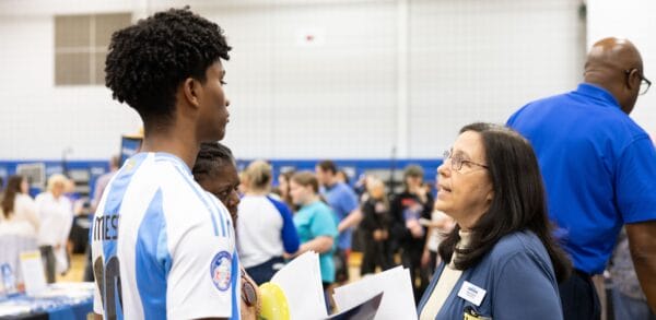 A young man in a soccer jersey speaks with a woman holding papers at the SUNY Niagara Open House, surrounded by several people and informational tables at a busy indoor event.