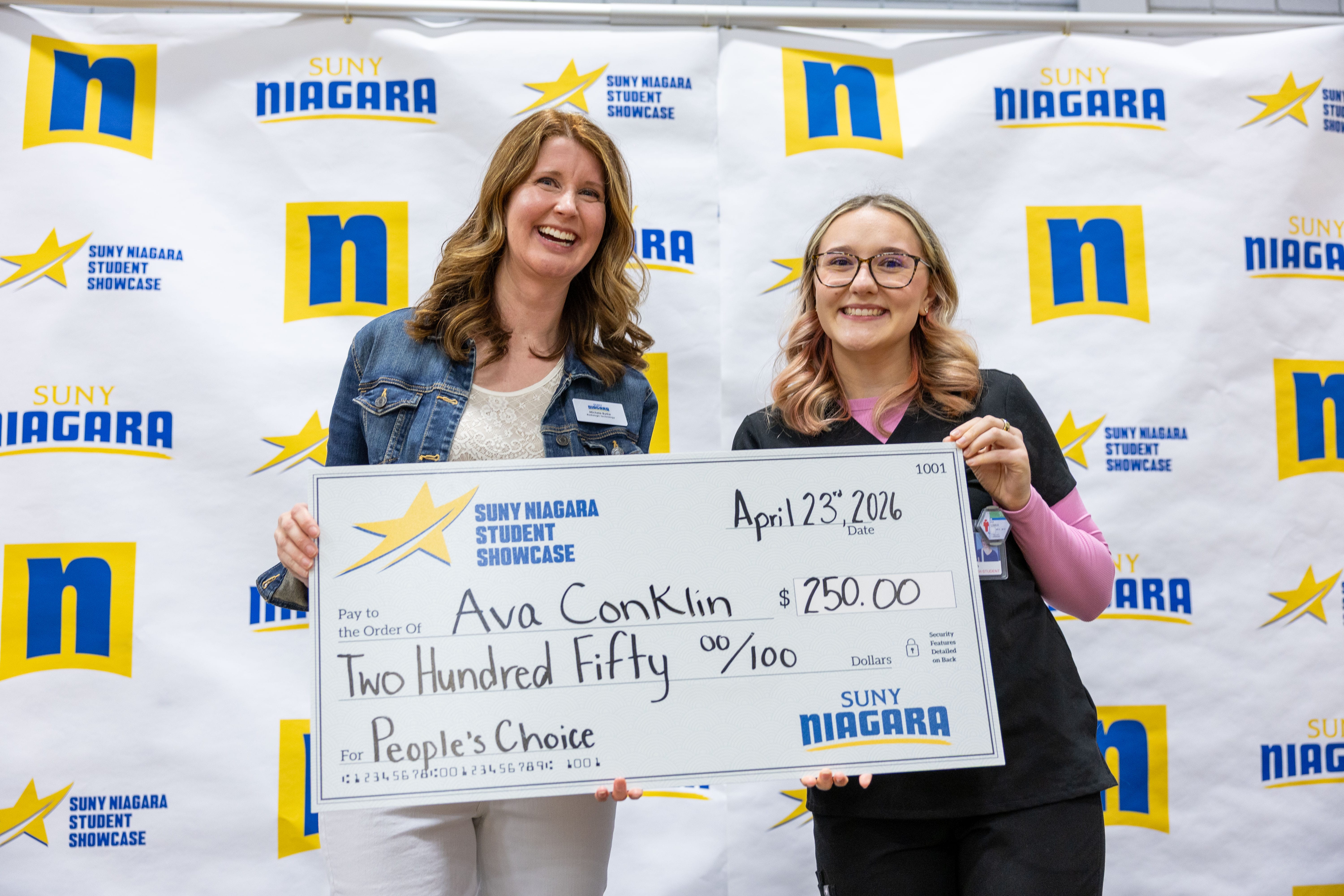 Two women smile while holding a large check for $250 made out to Ava Conklin for winning the “People’s Choice” award at the SUNY Niagara Student Showcase. A SUNY Niagara step-and-repeat banner is in the background.