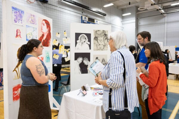Several people stand and talk at an indoor art exhibit, with sketch portraits and colorful artworks displayed on white panels. Art supplies and brochures are laid out on a table between them.