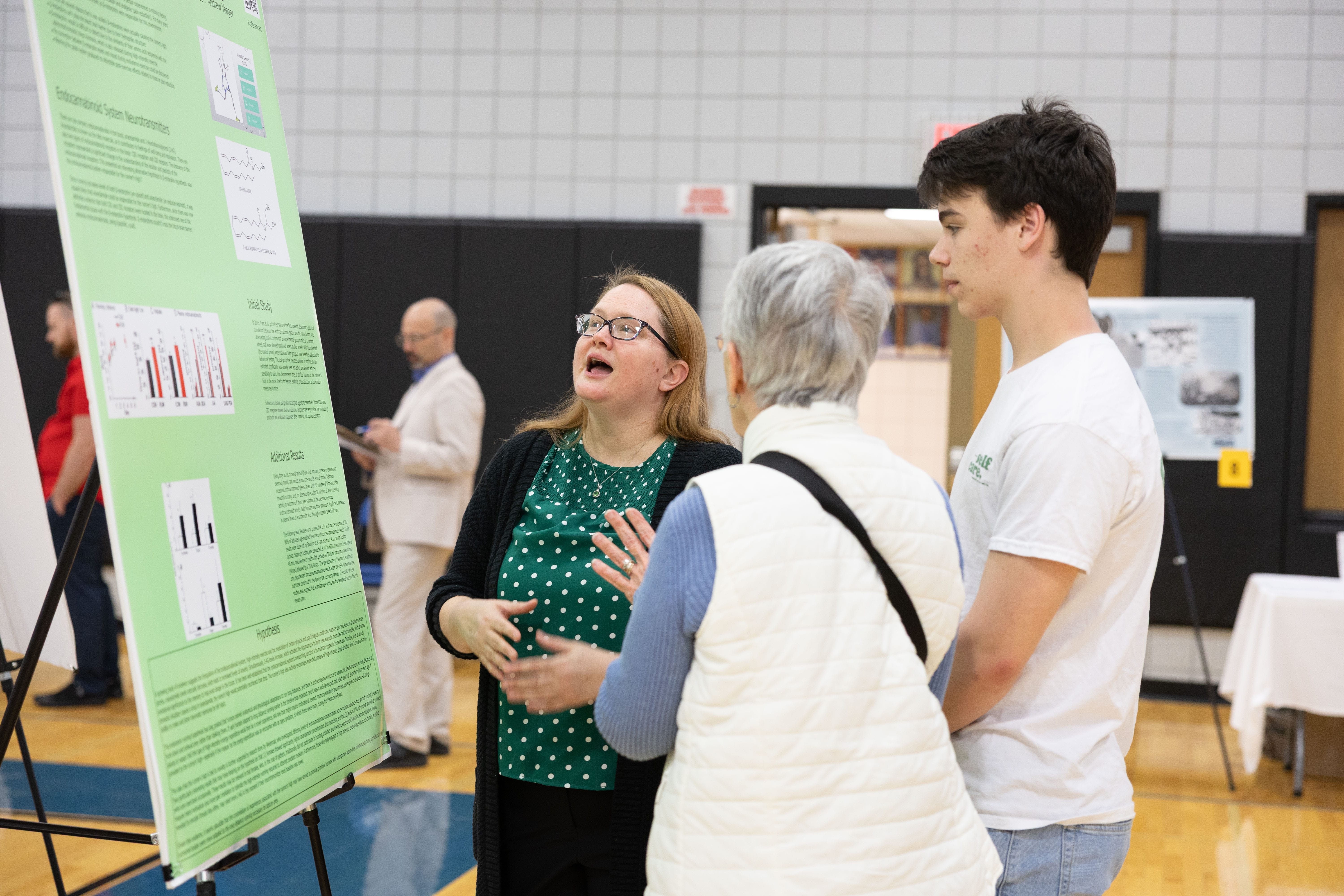 A woman enthusiastically explains a scientific poster to an older woman and a young man in a gymnasium during a science fair or academic event. Other people are visible in the background.