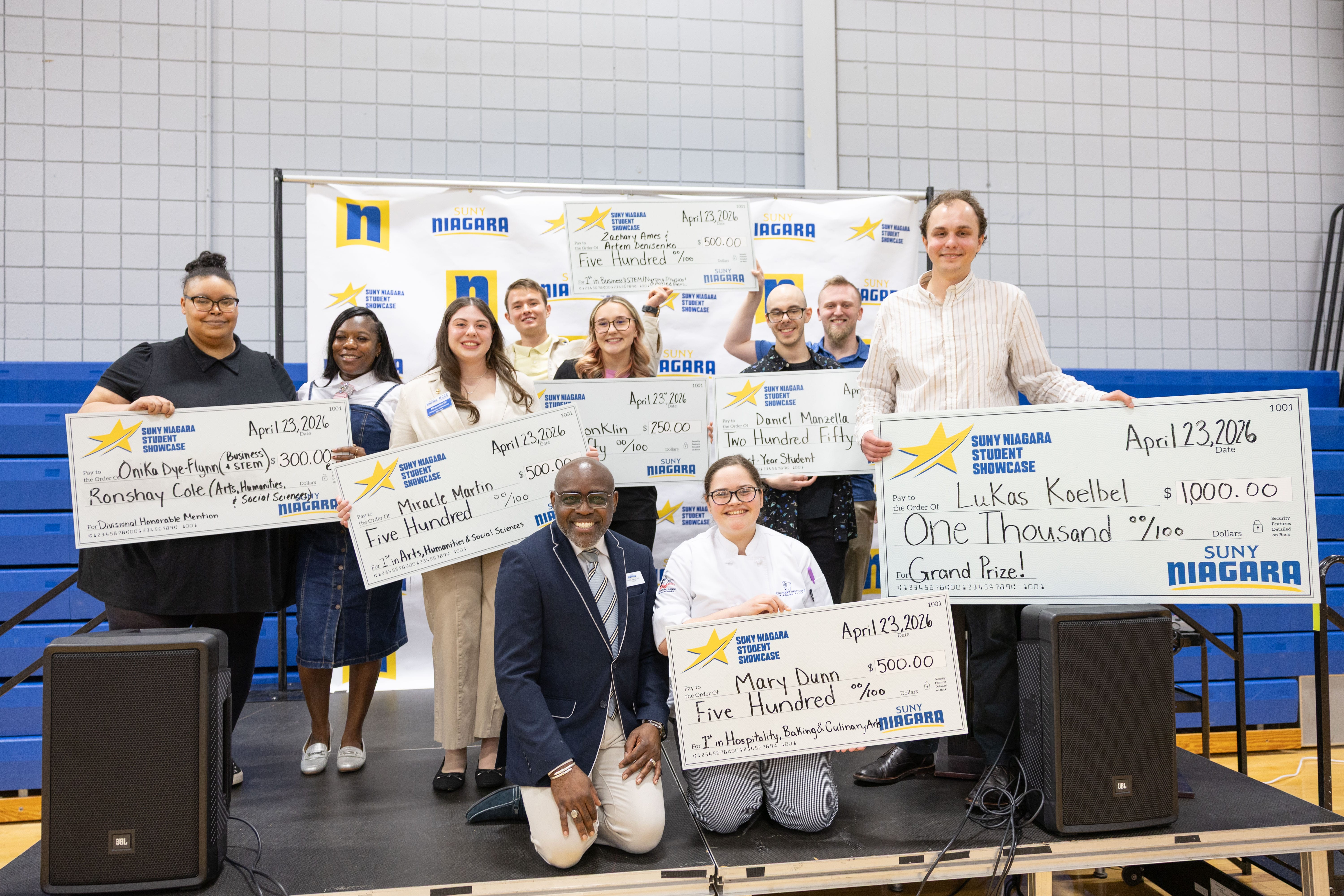 A group of people on a stage smile and hold oversized checks for various prize amounts. A branded backdrop featuring SUNY Niagara is behind them. Some are standing and others are kneeling in front.