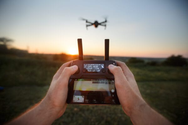 Hands holding a drone controller with a screen, flying a drone outdoors at sunset over a grassy field. The drone is visible in the sky, with the sun low on the horizon in the background.