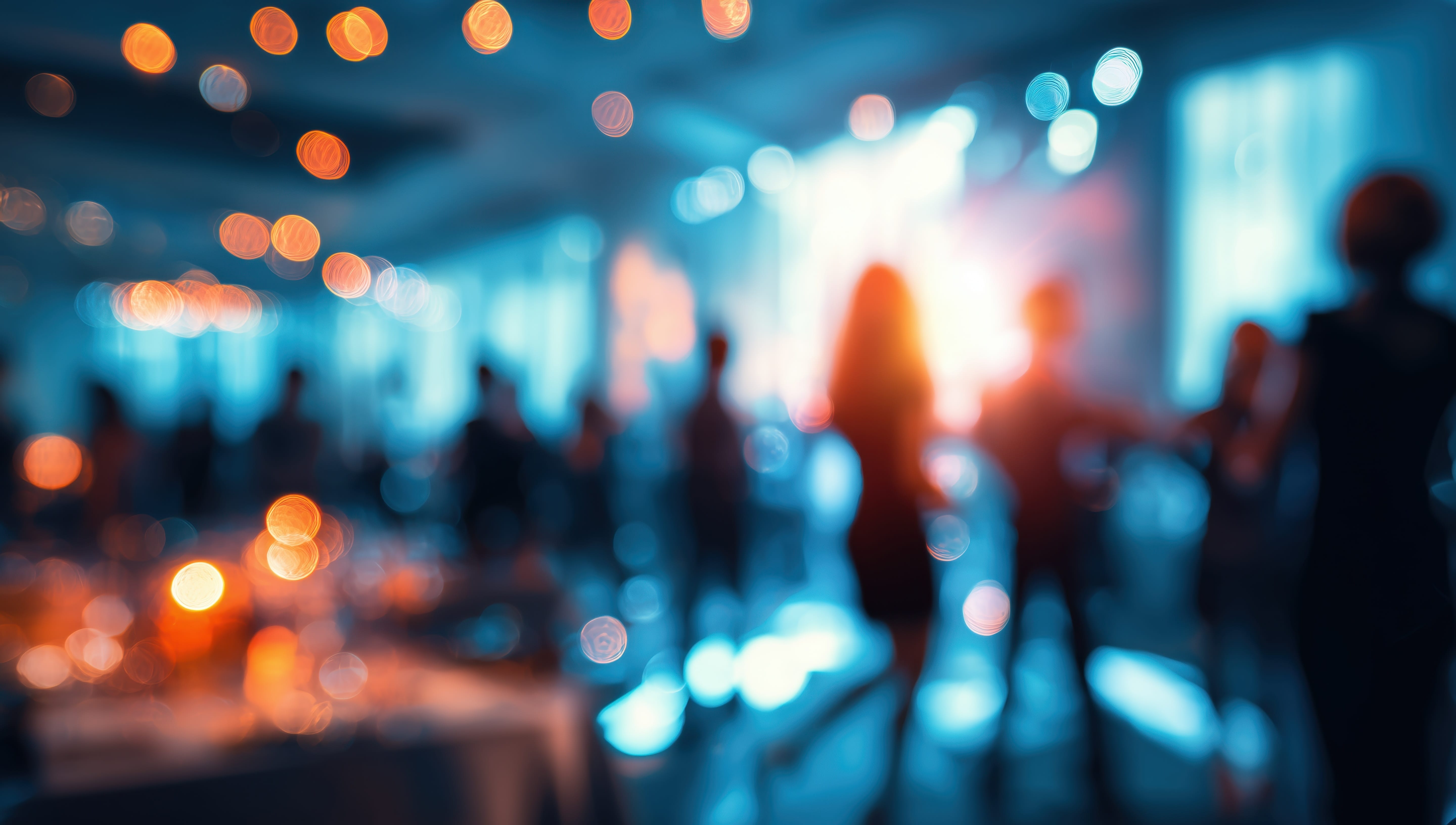 A blurred image of people dancing and mingling at an indoor event or party, with warm lights and colorful bokeh creating a festive and lively atmosphere.