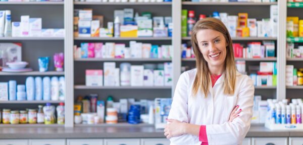 A pharmacist in a white coat stands with arms crossed and smiles in front of shelves stocked with various medicines and pharmaceutical products in a pharmacy.