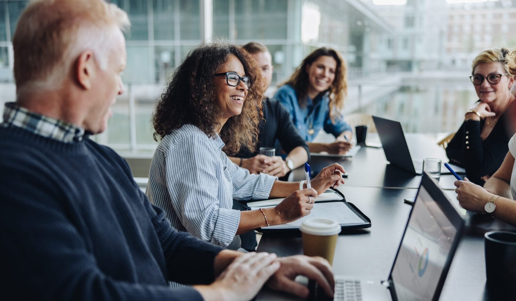A group of six people sit around a conference table with laptops, notebooks, and coffee cups, engaged in a lively discussion in a modern, glass-walled office.