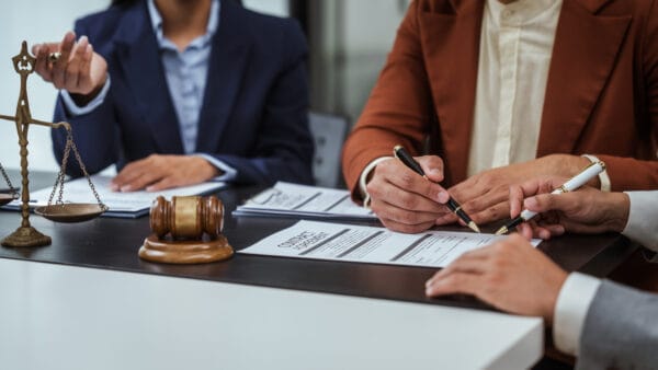 Three people in business attire sit at a table with legal documents, a gavel, and scales of justice, discussing and signing contract agreements in a professional office setting.