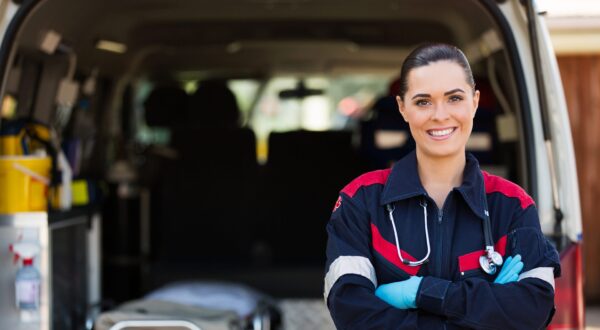 A smiling paramedic stands with arms crossed in front of an open ambulance, wearing a dark uniform, blue gloves, and a stethoscope around her neck. Medical equipment and a stretcher are visible inside the vehicle.