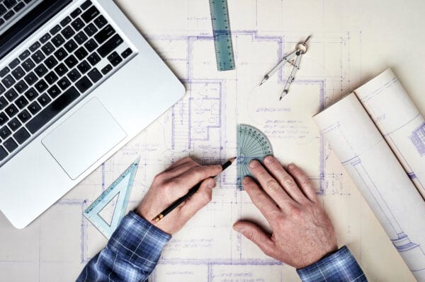 A person uses a protractor and pencil on architectural blueprints, with a laptop, ruler, compass, and rolled-up plans nearby on the desk.