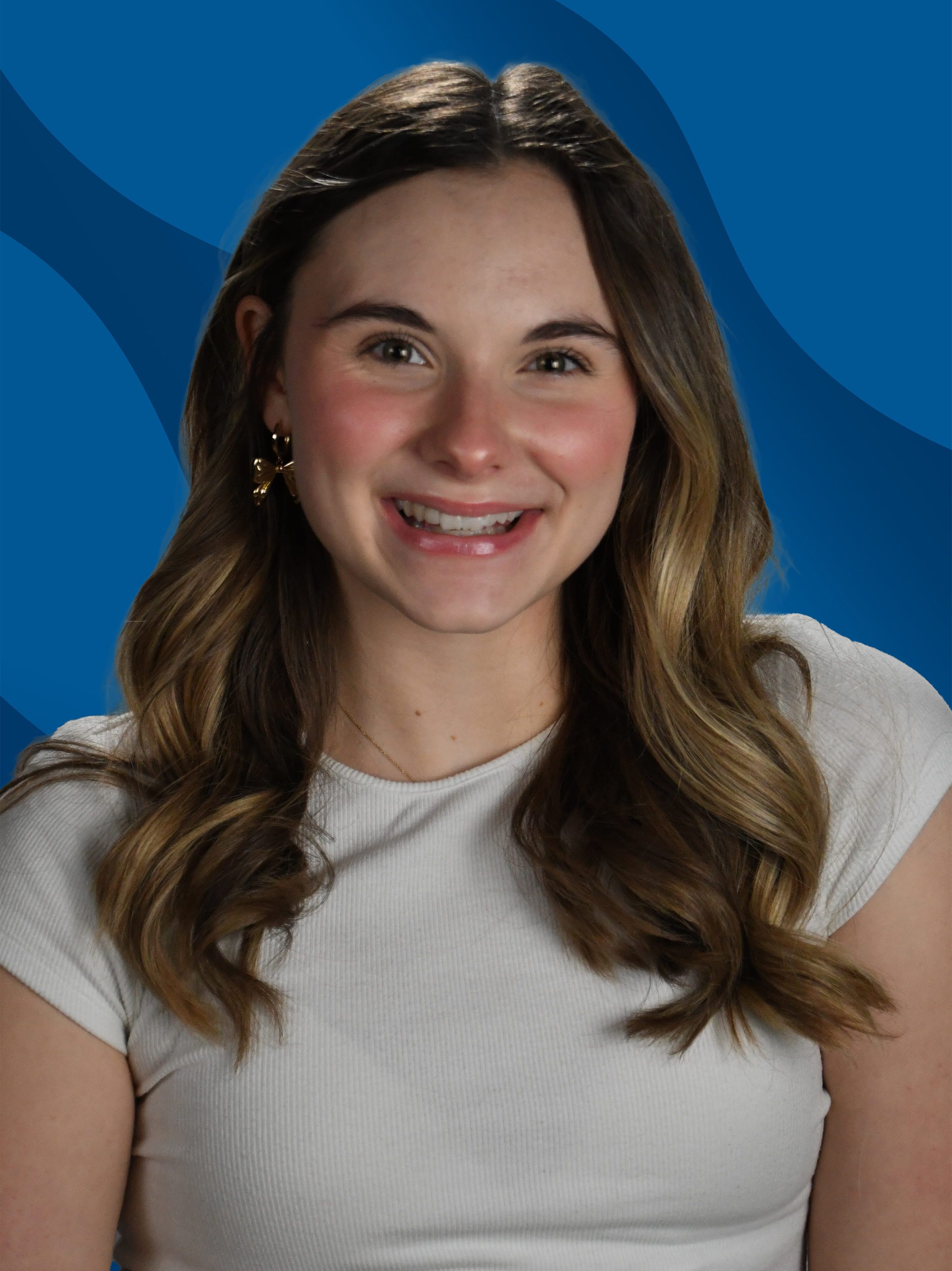 A young woman with wavy brown hair, wearing a short-sleeved white top, smiles in front of a blue abstract background.