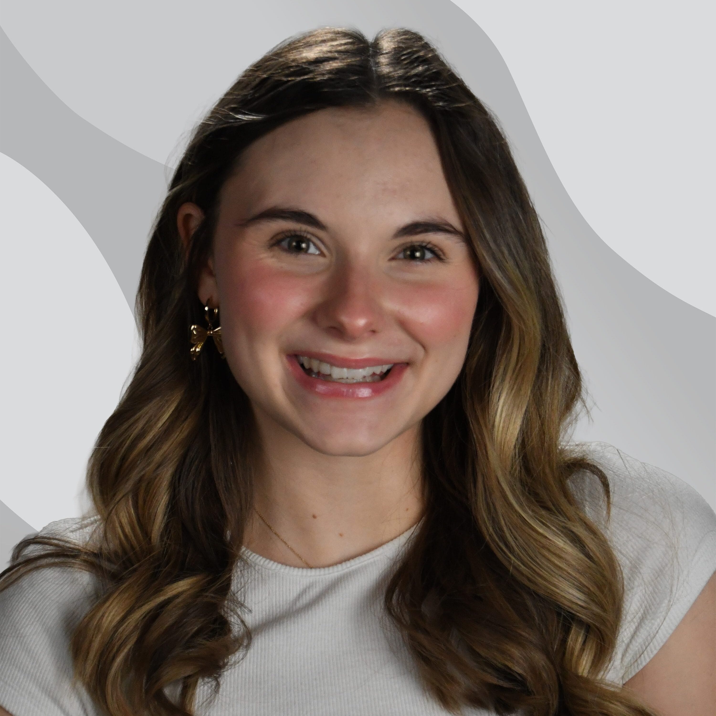 A smiling young woman with wavy brown hair wearing a white short-sleeve top, gold earrings, and a light gray abstract background.