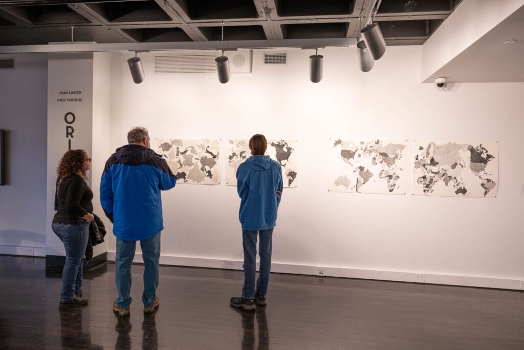 Three people stand in a gallery, looking at a series of world maps displayed on a white wall under spotlights. The floor is dark, and the ceiling shows exposed pipes and lights.