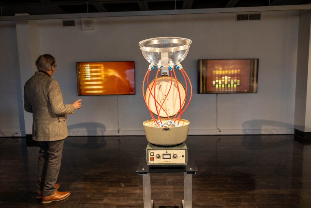 A person stands near an electronic art installation with red tubes and a large clear bowl on top, displayed on a black pedestal. Two digital screens with abstract yellow graphics are mounted on the wall in the background.
