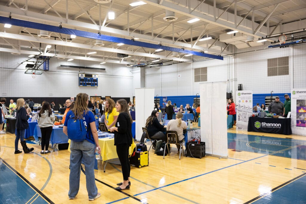 A gymnasium filled with people attending a job or college fair. Booths with banners and tables are set up around the gym, with attendees interacting with representatives. The atmosphere is busy and well-lit.