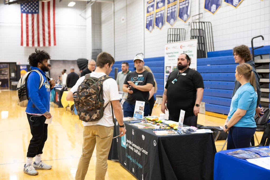Students with backpacks talk to representatives at an informational booth in a gymnasium, with blue bleachers, banners, and a large American flag visible in the background. Pamphlets and snacks are on the table.
