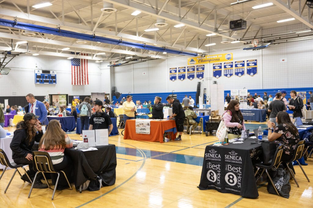 People sit and talk at tables covered with informational materials in a gymnasium during a college or job fair. Banners, an American flag, and blue walls are visible in the background.