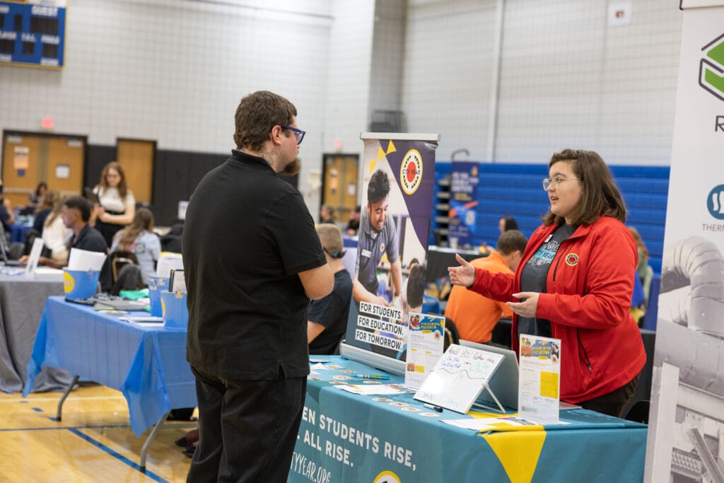 A woman in a red jacket speaks to a man at an information booth during a career fair in a gymnasium, with other tables and people visible in the background.