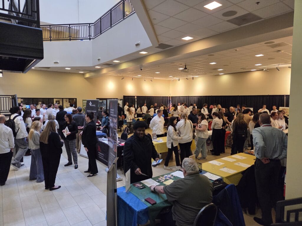 A large group of people gather at an indoor event with various tables, informational booths, and displays. Attendees are interacting and networking in a spacious, well-lit hall.