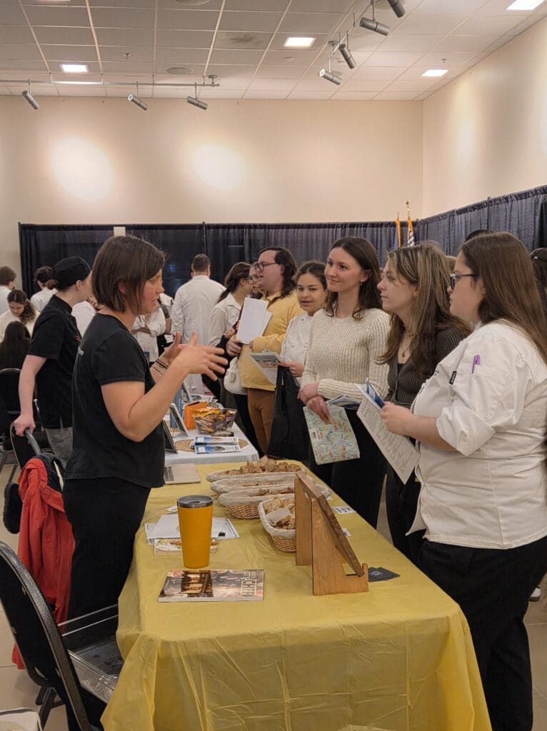 A woman stands behind a yellow-covered table speaking to a group of people, some in white chef coats, at an indoor event or fair with booths and other attendees in the background.