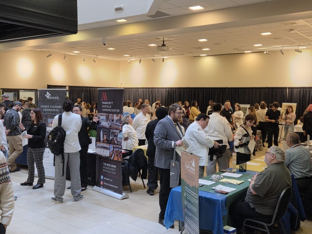 A large group of people interact at a busy indoor career fair, with tables covered in brochures and banners for hospitality and culinary companies. Many attendees wear white chef jackets, and the room is well-lit with overhead lights.