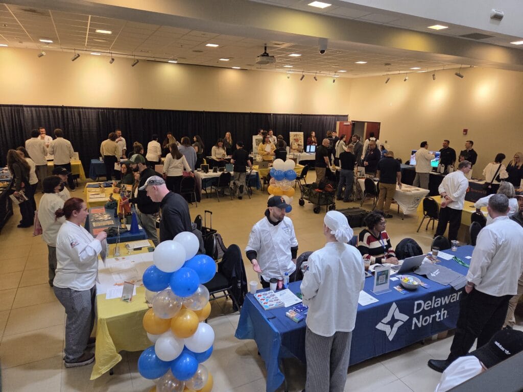 A busy job fair or expo in a large indoor space, with people interacting at various decorated tables, some wearing chef coats and hats. Blue, white, and gold balloons are near the Delaware North booth in the foreground.