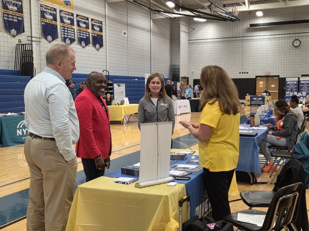 Four adults stand and talk around a table covered with a yellow cloth at a college fair in a gymnasium, with informational booths and banners visible in the background.