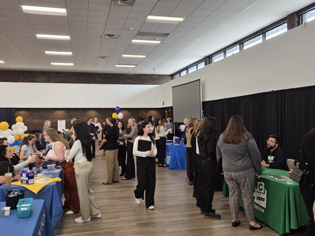 A large group of people interacts at an indoor career fair, visiting tables with informational materials and balloons. Several booths are set up around the room, and attendees are engaged in conversation.