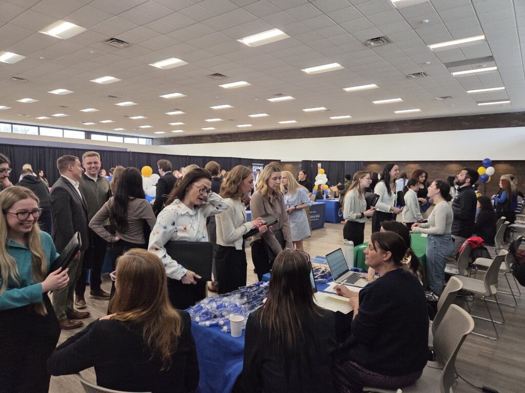 Large room with people standing in lines and talking at tables covered with blue cloths. Attendees hold folders and chat with seated representatives; some tables have giveaways. Overhead lights and balloons are visible.