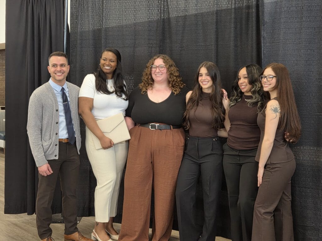 Six people stand together, posing and smiling in front of a black curtain. Five women and one man are dressed in business casual attire, with mostly brown, black, and white clothing.