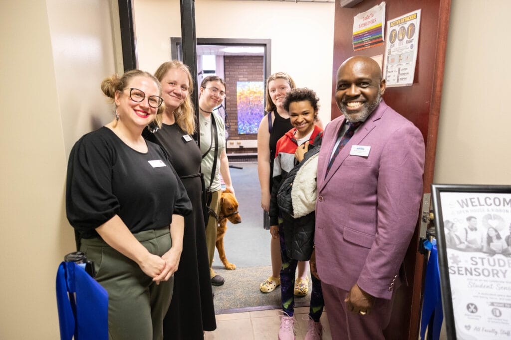A group of six people, including one staff, students and a service dog, smile at the entrance of a room. Four adults wear name tags, and one man in a mauve suit stands near a welcome sign for a student sensory room.