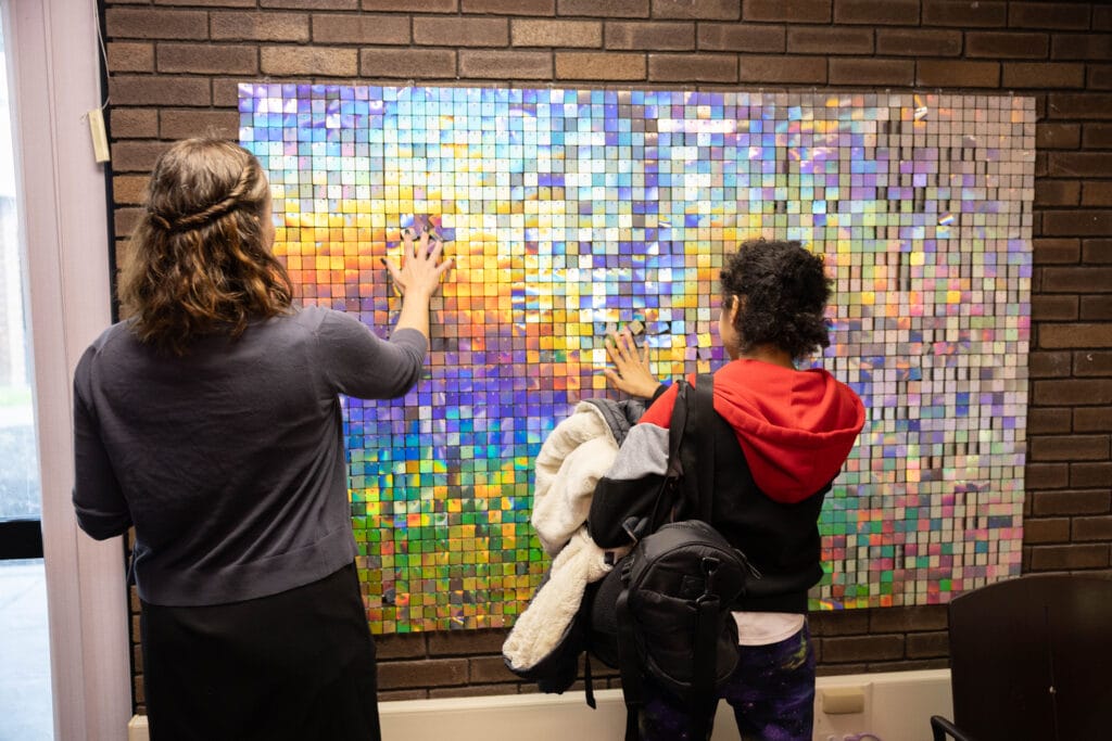 Two people stand facing a colorful, iridescent sequin art panel on a brick wall, touching the surface with their hands. One person holds a jacket and backpack.
