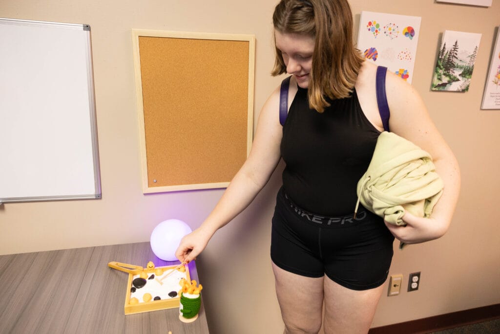 A young woman in a black outfit holds clothes in one arm and uses a wooden grabber to pick up objects from a sensory box on a table in a decorated room.