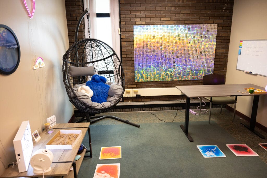 A cozy sensory room with a hanging egg chair, plush toys, a mosaic wall art, a sandbox table, colorful floor mats, a desk, and a whiteboard, creating a calm and inviting atmosphere.