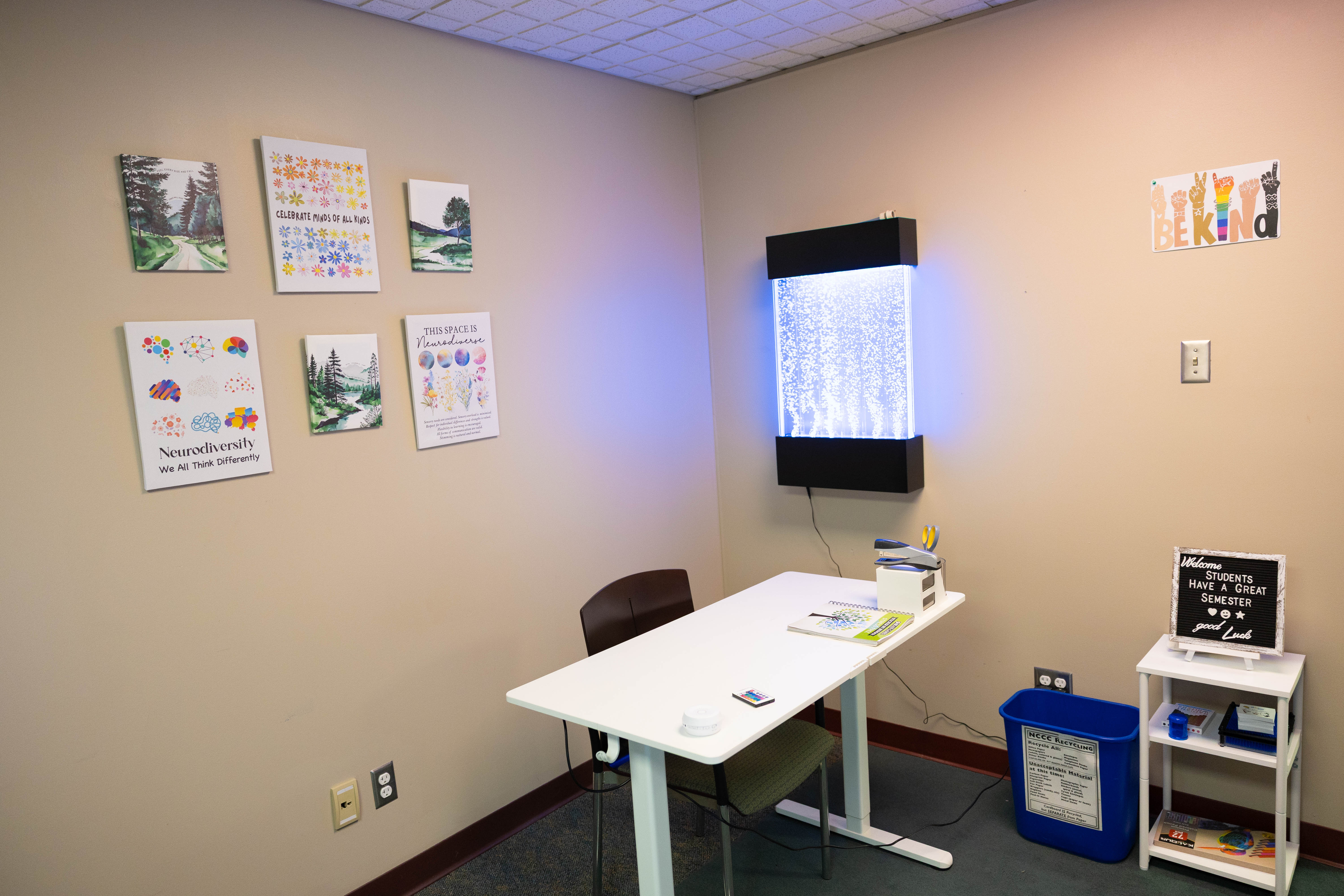 A small office with a white desk and chair, a bubbling wall panel, motivational posters and artwork on the beige walls, and a blue recycling bin beside the desk.