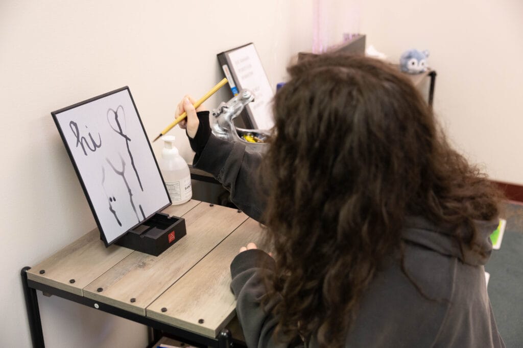 A person with long hair uses a brush to paint “hi” and a heart on a small upright board at a wooden desk. A bottle and small items are on the desk, and shelves with plush toys are in the background.
