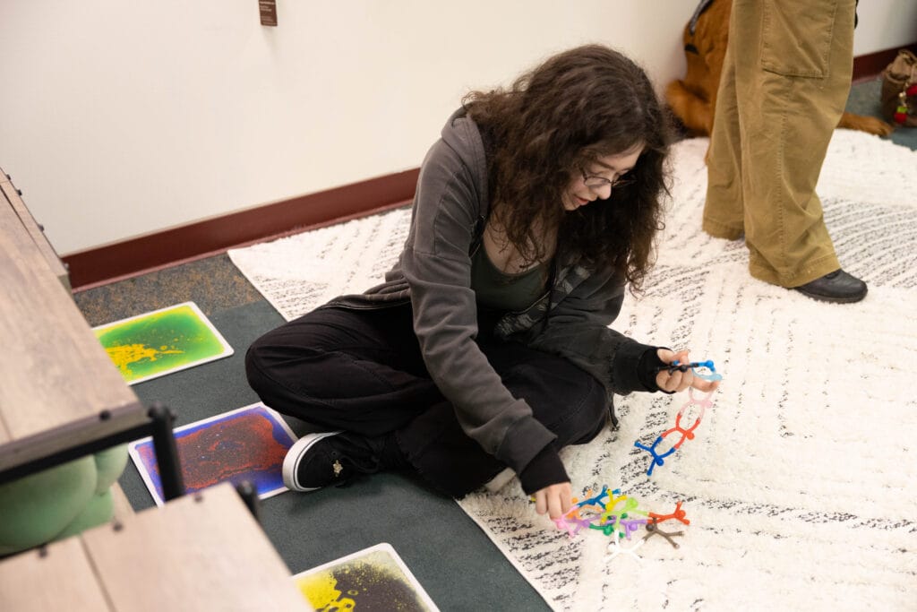 A young person with long curly hair sits cross-legged on a rug, playing with colorful toys. There are textured sensory pads on the floor nearby and another person standing in the background.