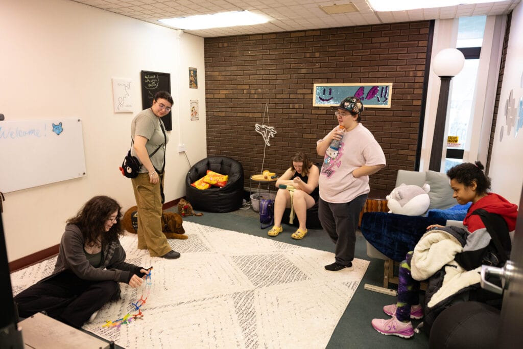 A group of five people relax in a cozy, casual room with bean bags, a whiteboard, and a brick wall. Some are sitting, one is standing, and they appear to be talking or playing with objects. The mood is relaxed and friendly.